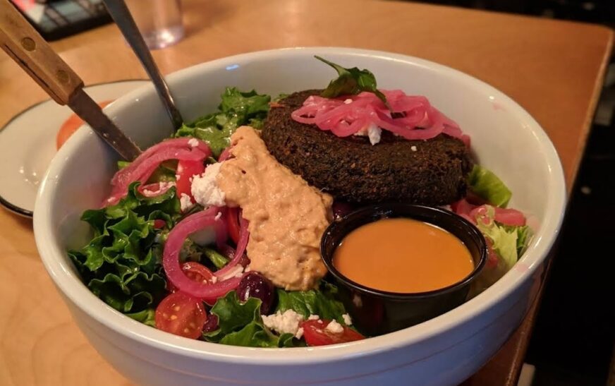 A vibrant falafel salad bowl served at Black Tap Craft Burgers & Beer in SoHo, featuring a crispy falafel patty on a bed of fresh greens, topped with pickled onions, vegan feta, hummus, and a side of dressing